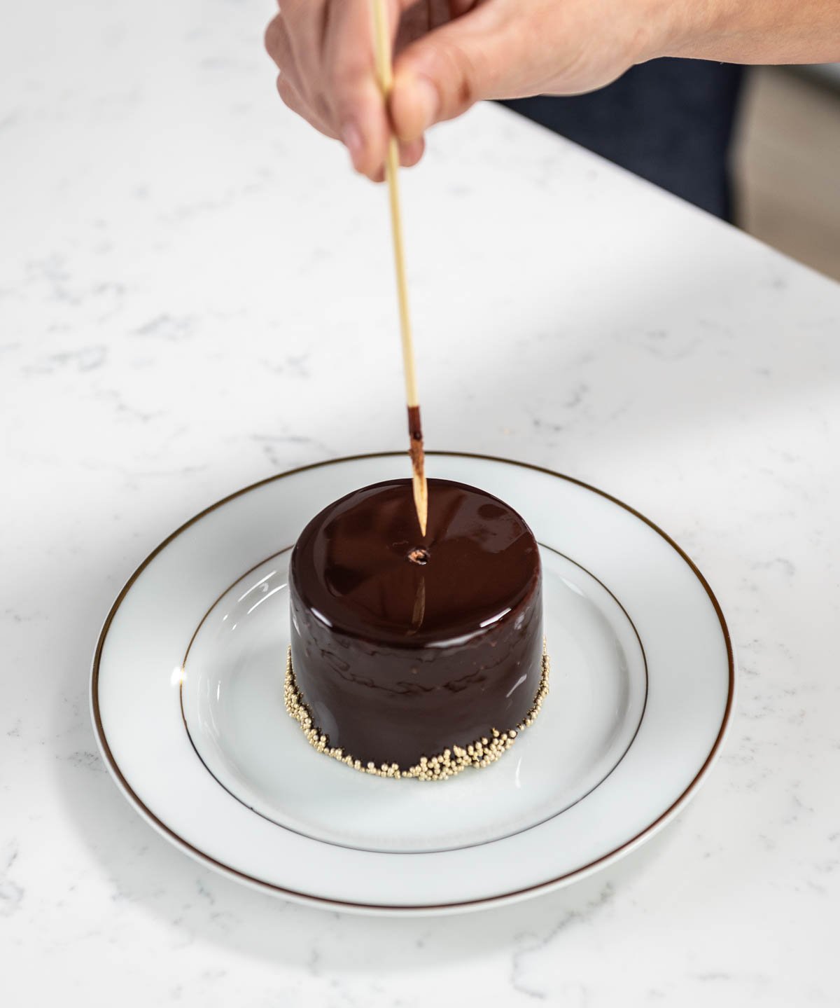 hand pulling toothpick out of hand placing edible gold on top of chocolate entremet