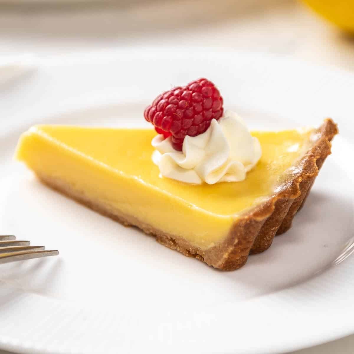 close up of a slice of lemon tart with creme fraiche and a raspberry on a white plate