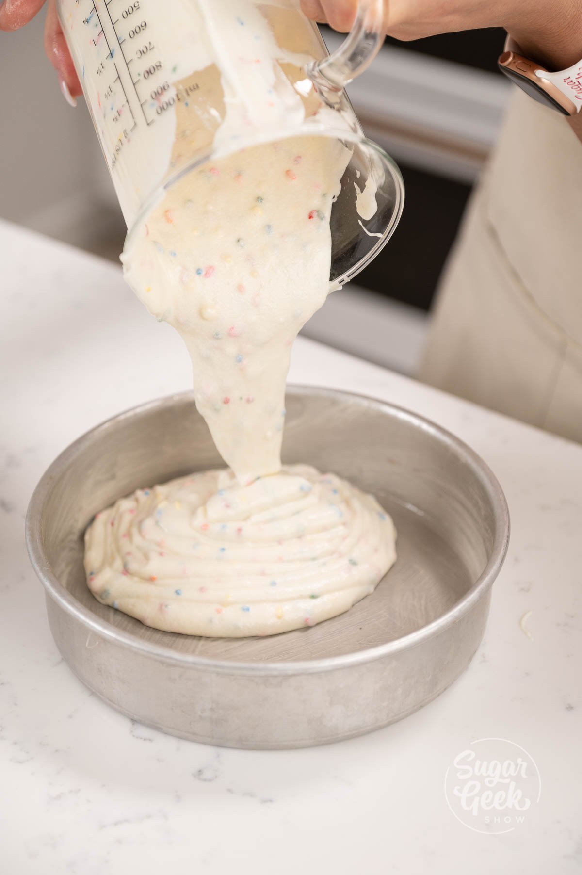 Glass measuring cup being held above cake pans pouring cake batter into the cake pans