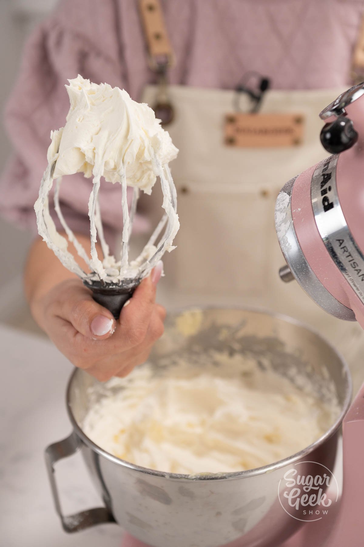 hand holding whisk covered in buttercream above mixing bowl.