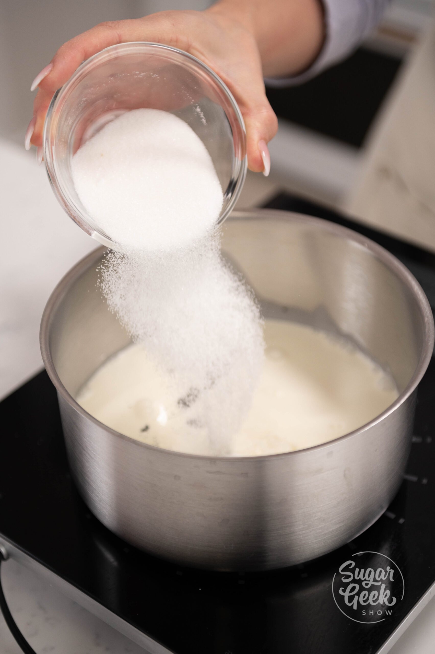 hand pouring bowl of sugar into pot of vanilla and cream