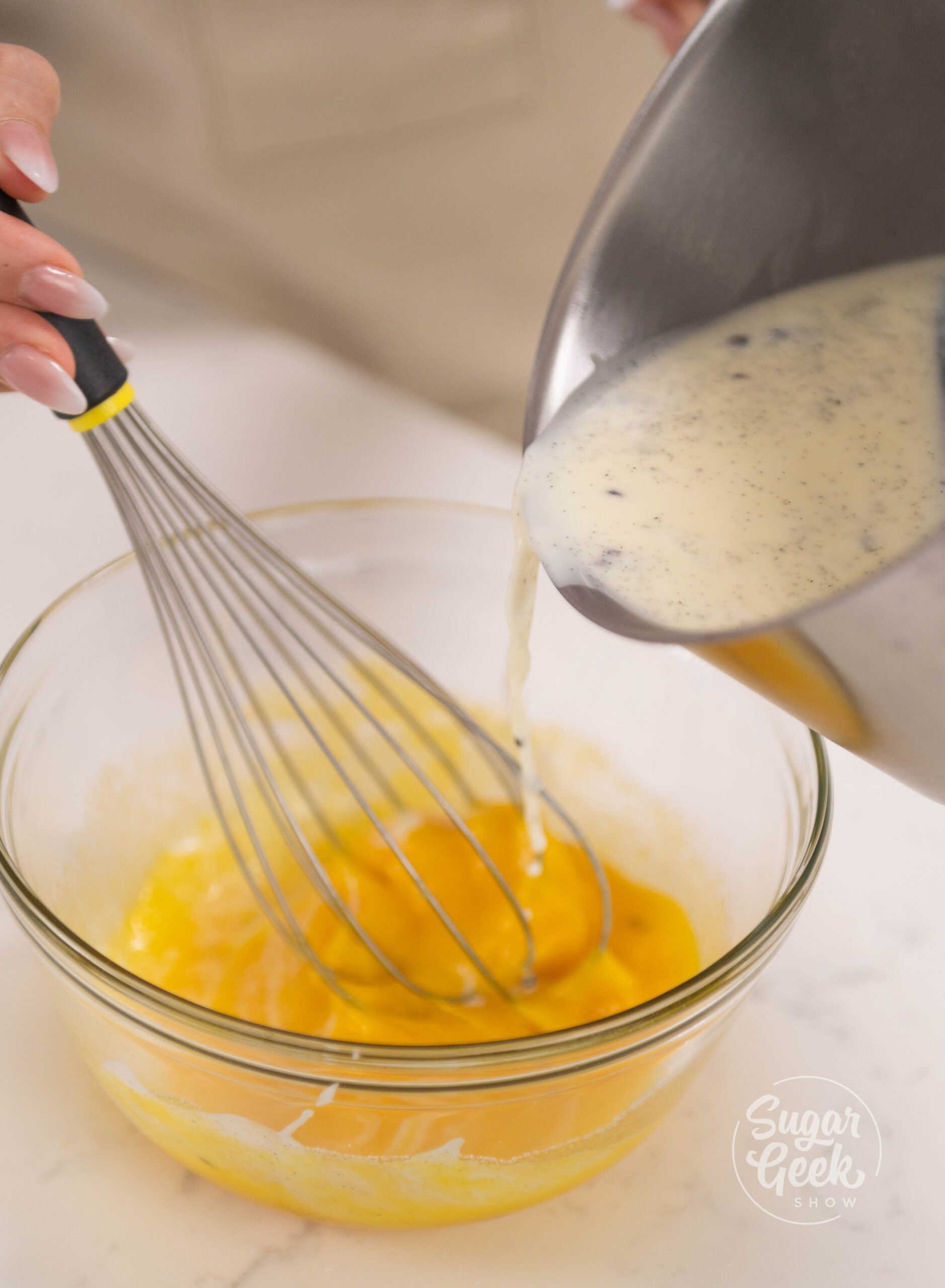 hand holding whisk and pouring bowl of cream into bowl off egg yolks