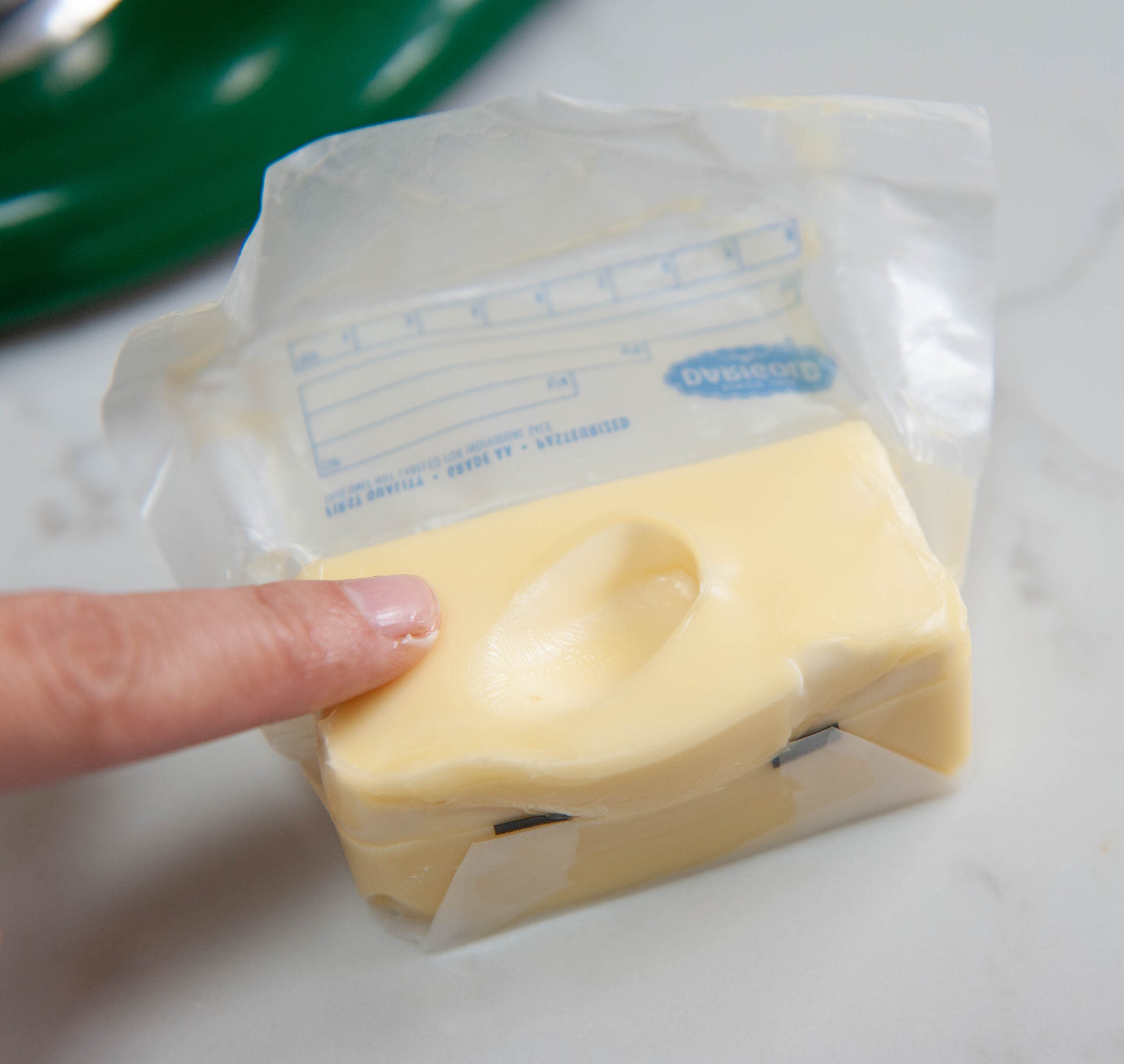 Finger pressing into a softened block of butter with its wrapper unwrapped on a white countertop.