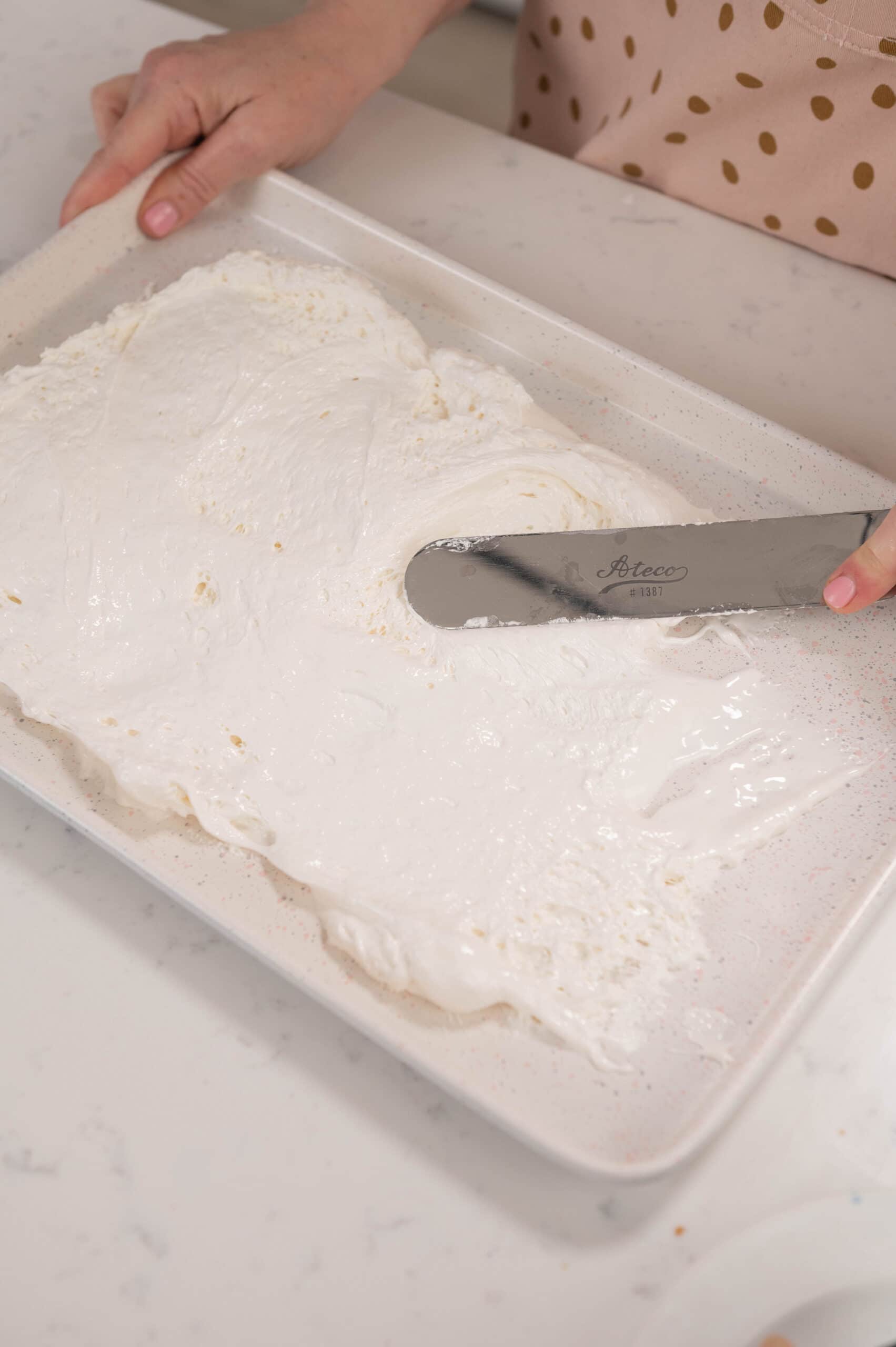 Hand using spatula to spread marshmallow fluff on to sheet pan.