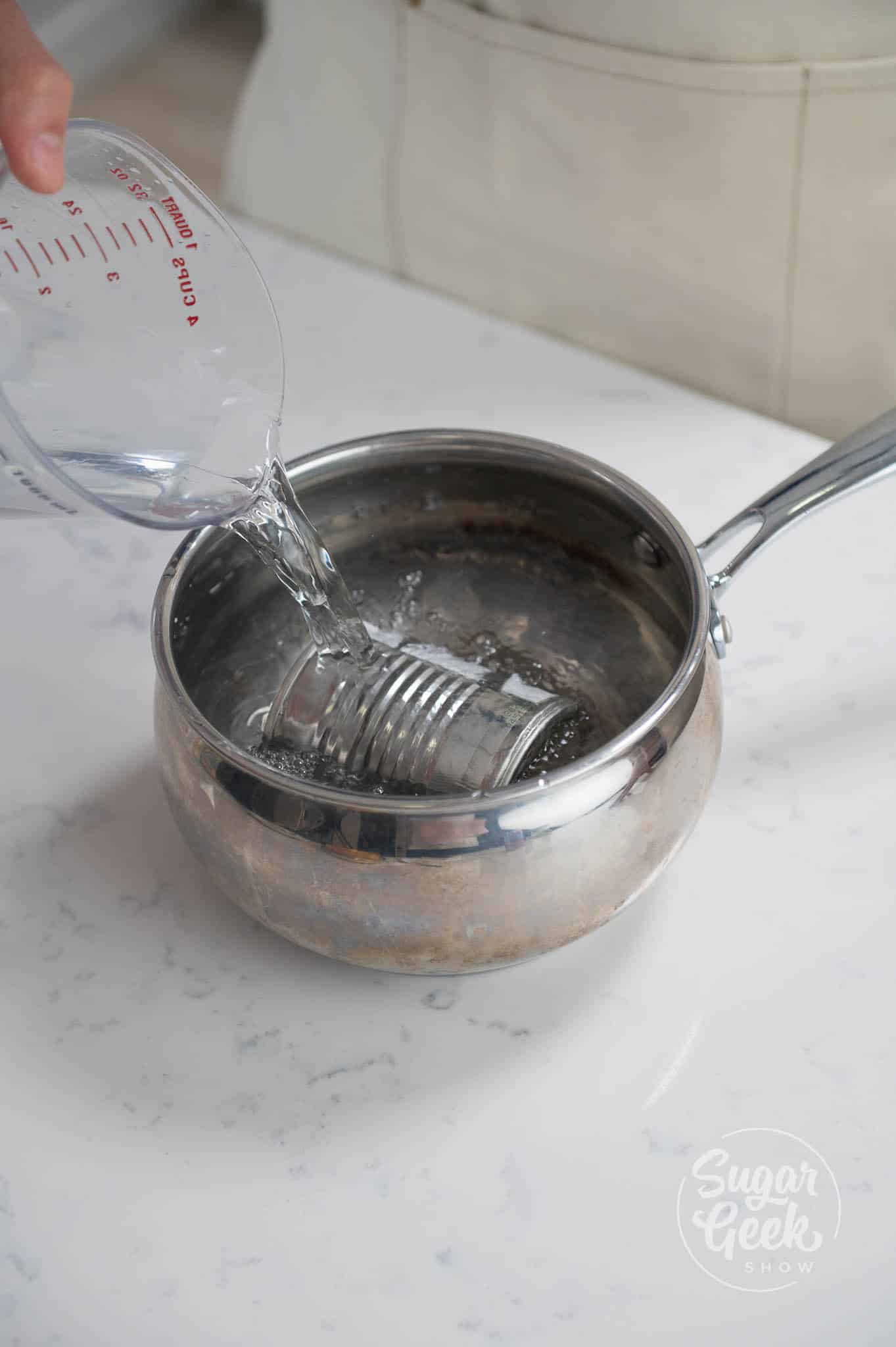 hand pouring measuring cup of water over pot with can inside.