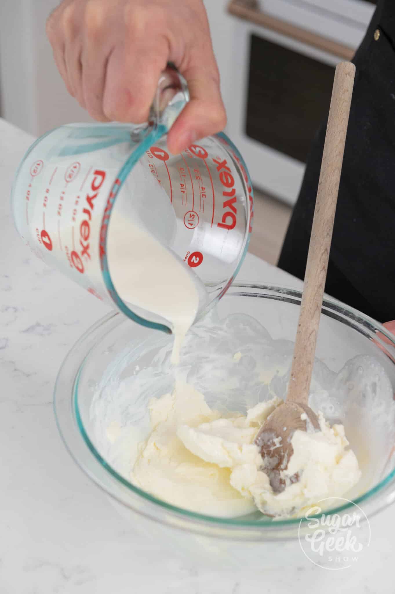 hand using measuring cup to pour cream onto cream cheese.