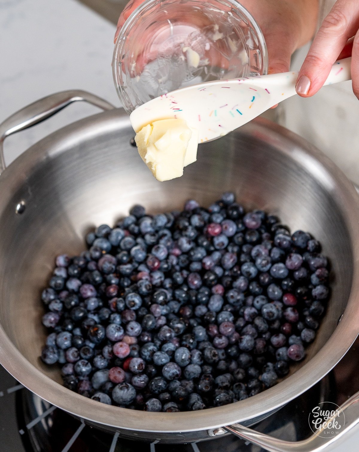 hand adding butter on a spatula to fresh blueberries in a pot.