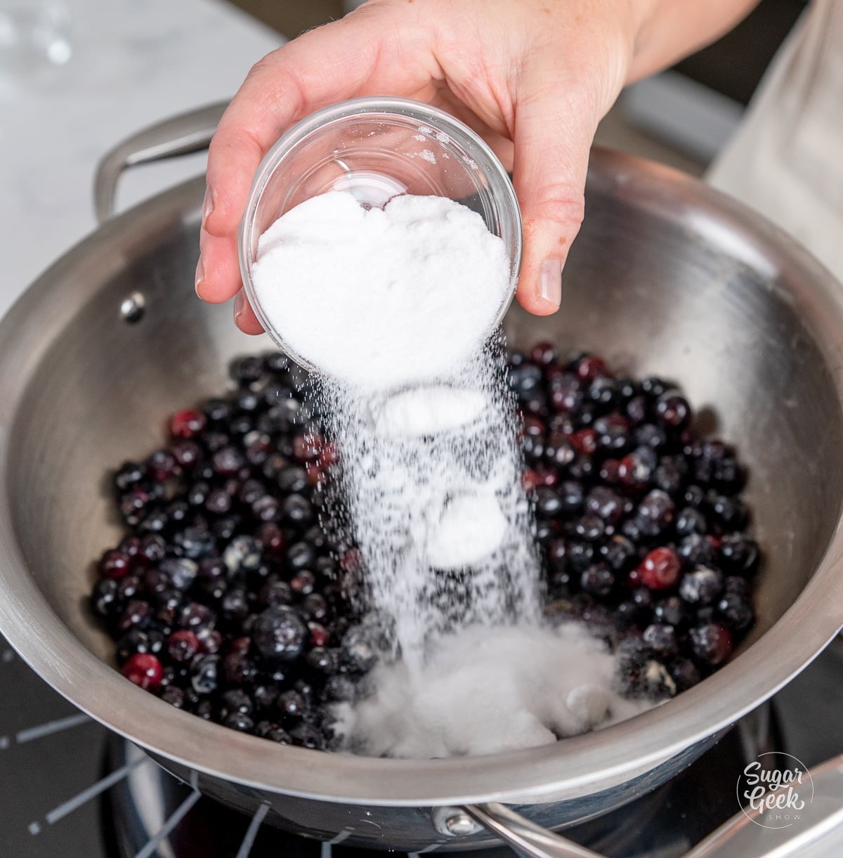 hand adding sugar to cooking blueberry mixture in a pot.