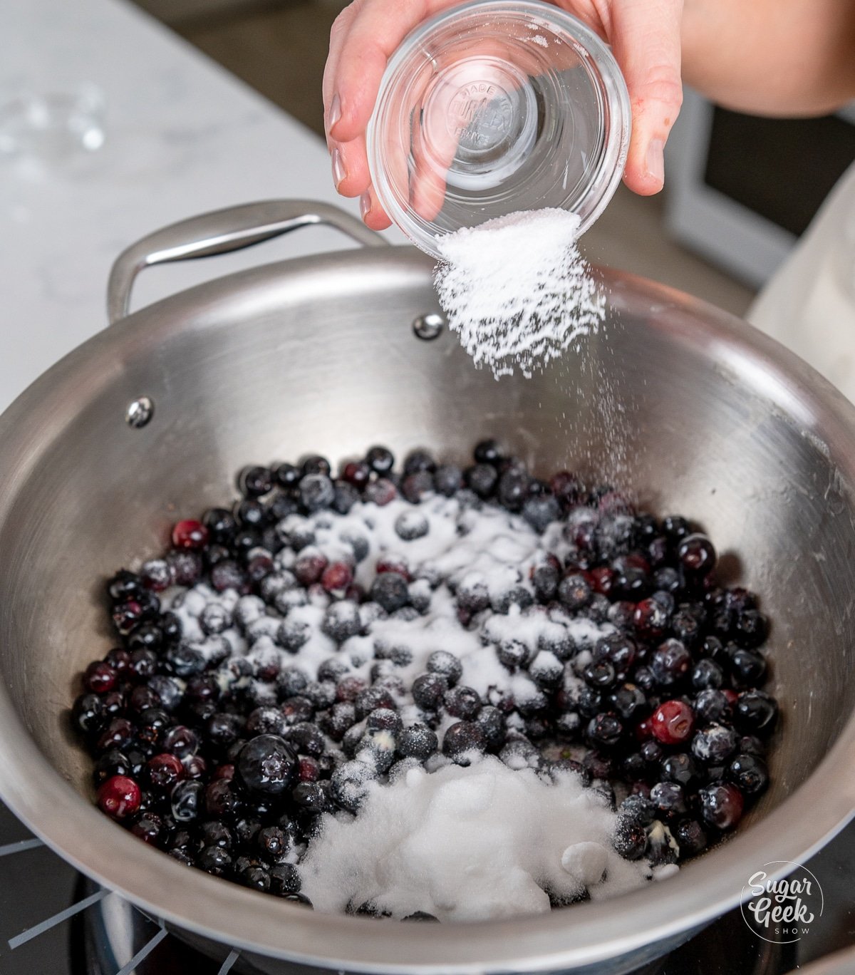 hand adding sugar to cooking blueberry mixture in a pot.