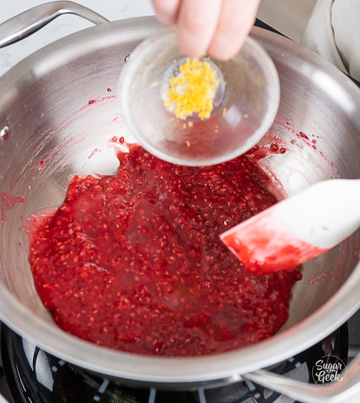 hand pouring lemon zest into pot with raspberry mixture.