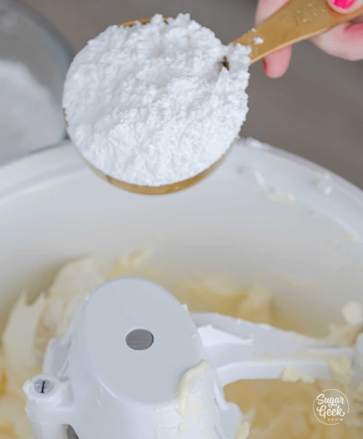 hand adding a measuring cup of powdered sugar to the mixing bowl. 