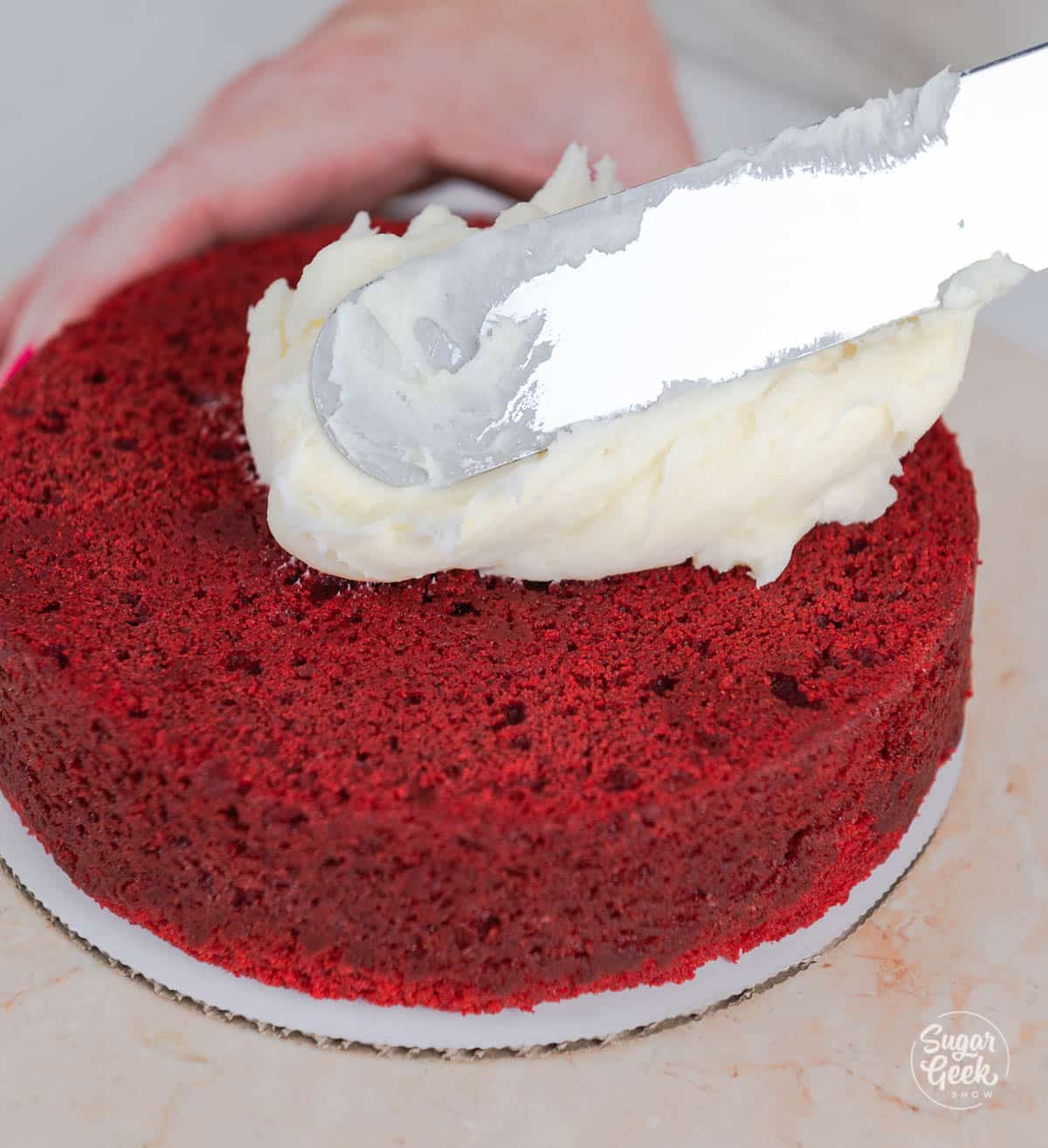 hands spreading cream cheese frosting on top of a layer of cake with a spatula.