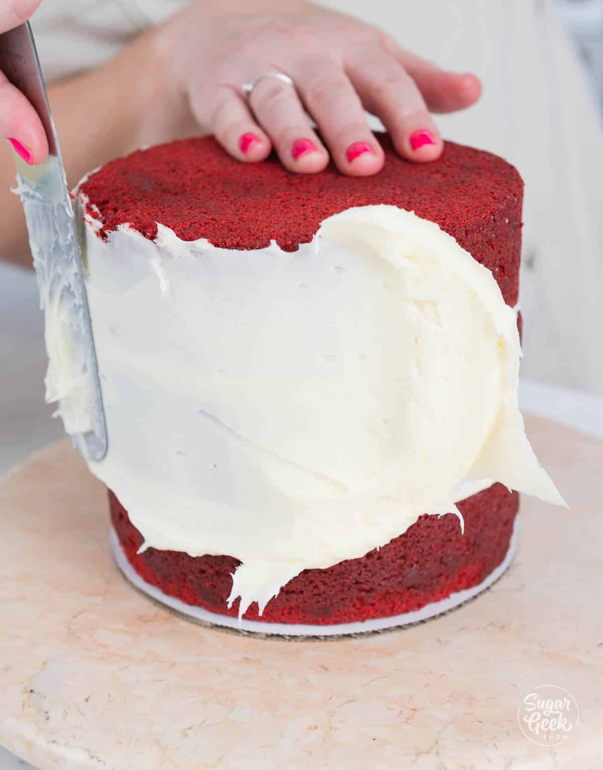 hands spreading a crumb coat of cream cheese frosting on the stacked cake. 