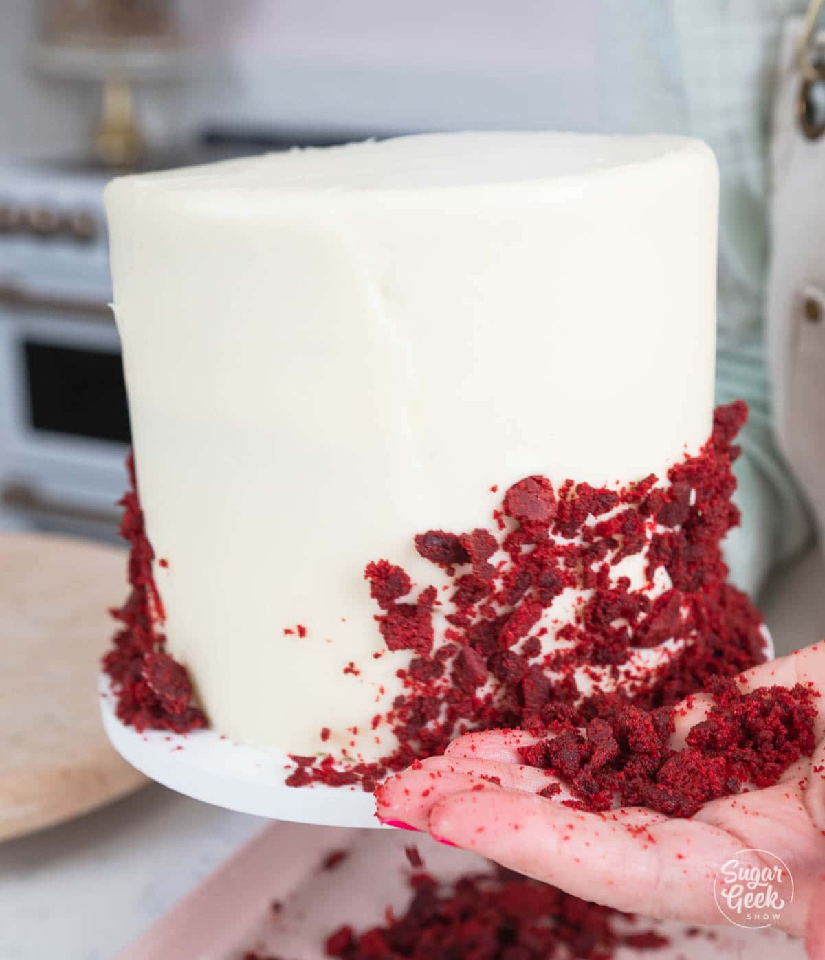hand pressing red velvet crumbs to the side of the frosted cake. 