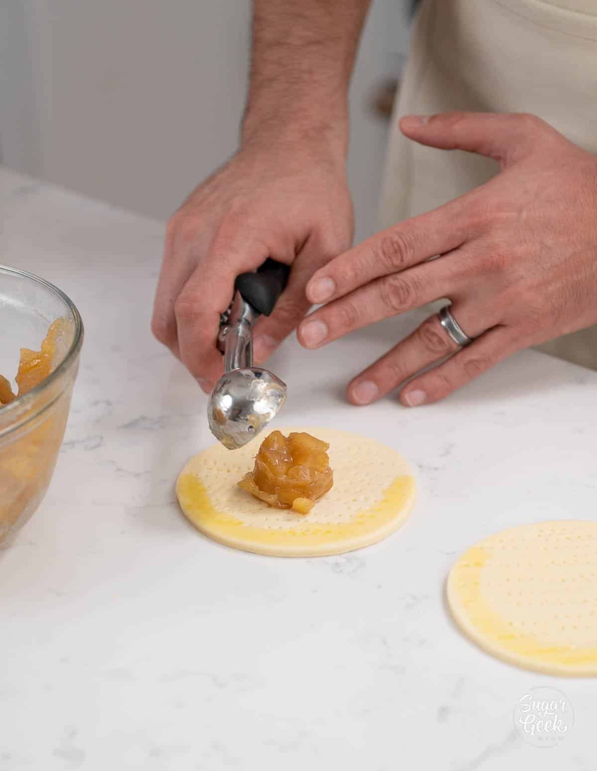 hand using a small scoop to scoop apple filling into center of dough. 