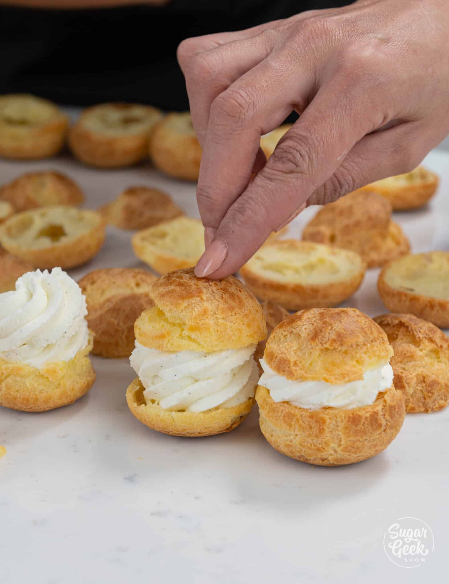 finger pushing the lid of a cream puff on top of whipped cream