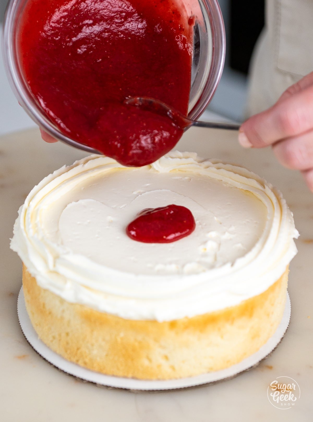hands pouring strawberry puree onto a cake layer with a buttercream dam. 