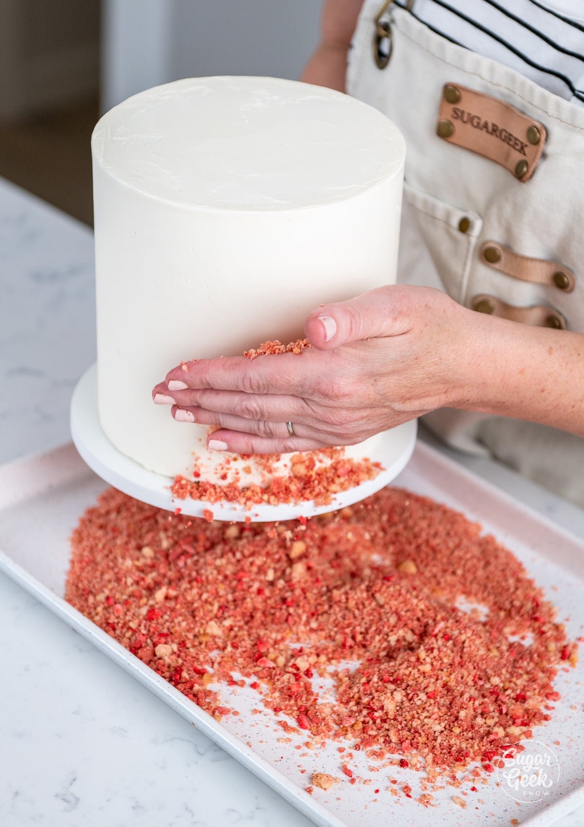 hands pressing strawberry crunch to the outside of the cake. 