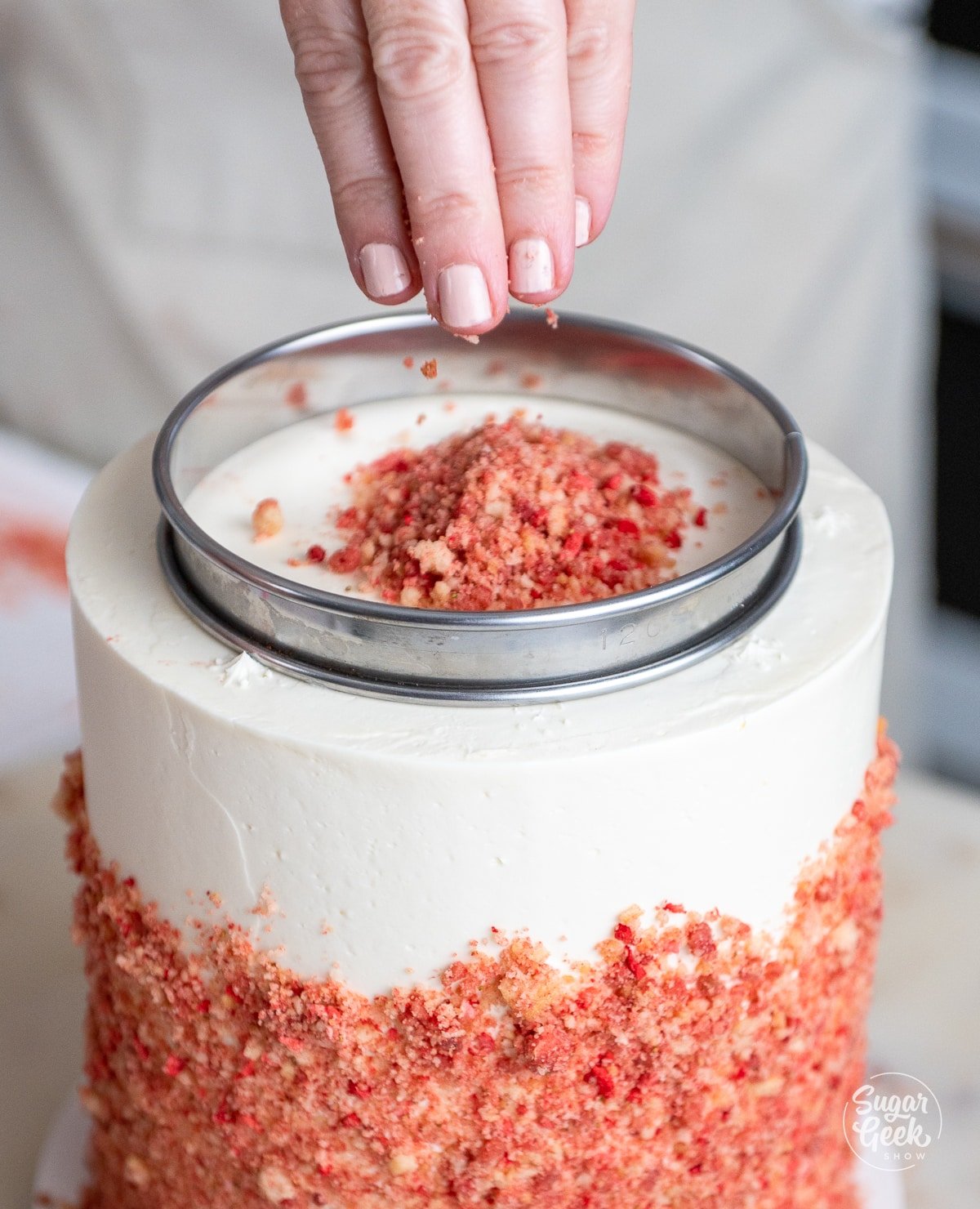 a hand sprinkling strawberry crunch into a ring on top of the cake. 