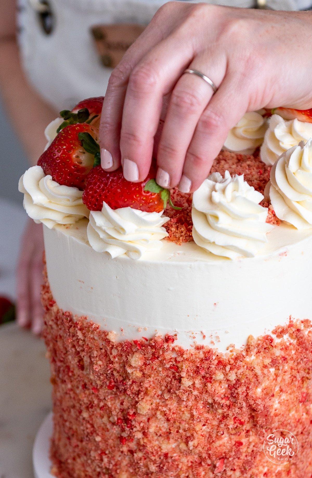 hands placing a strawberry onto buttercream dollops on top of the cake. 