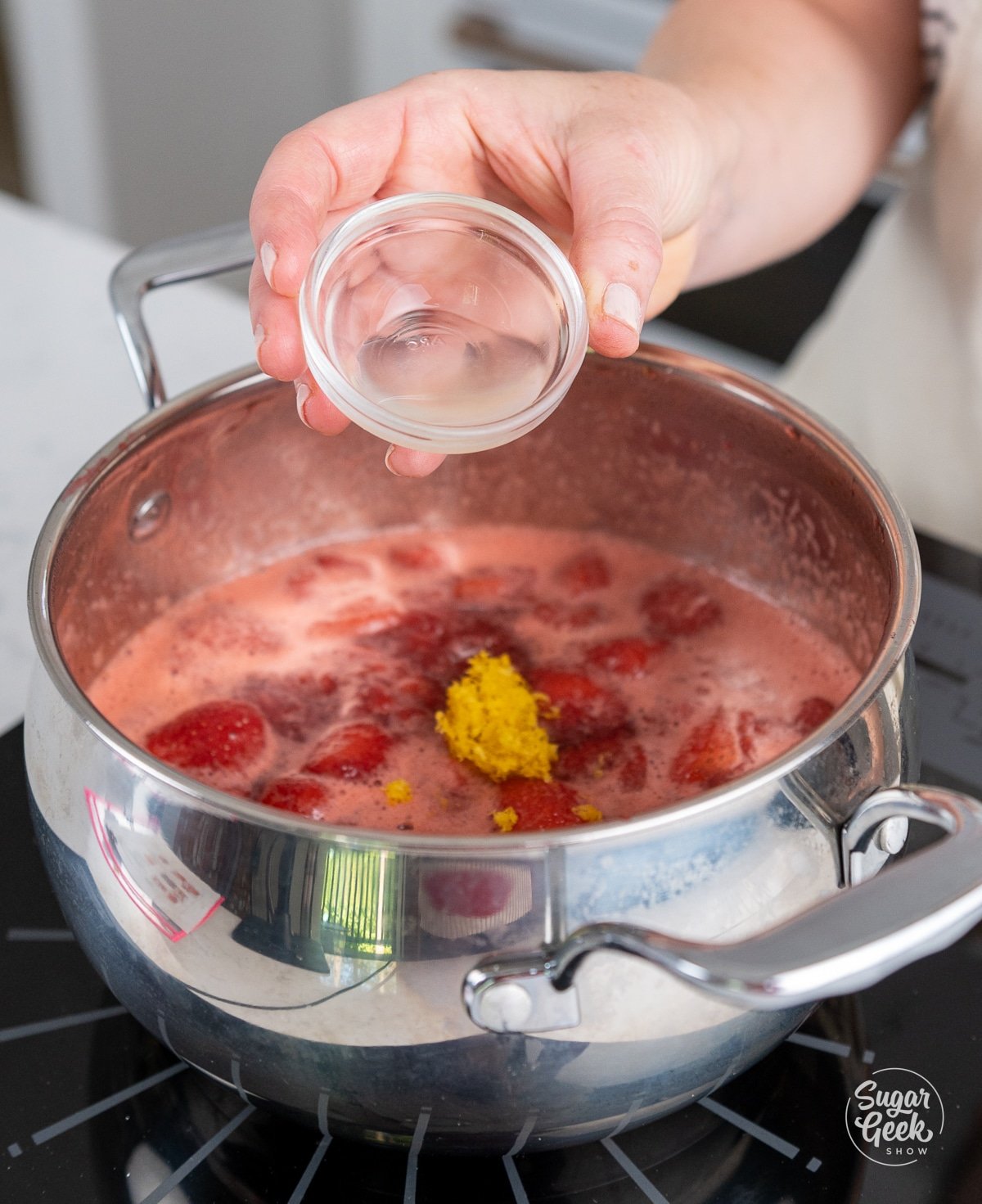 hand adding lemon extract to a pot with cooking strawberries and lemon zest.