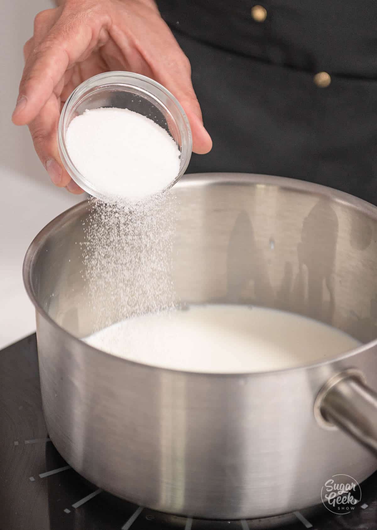 hand pouring sugar into saucepan with milk.