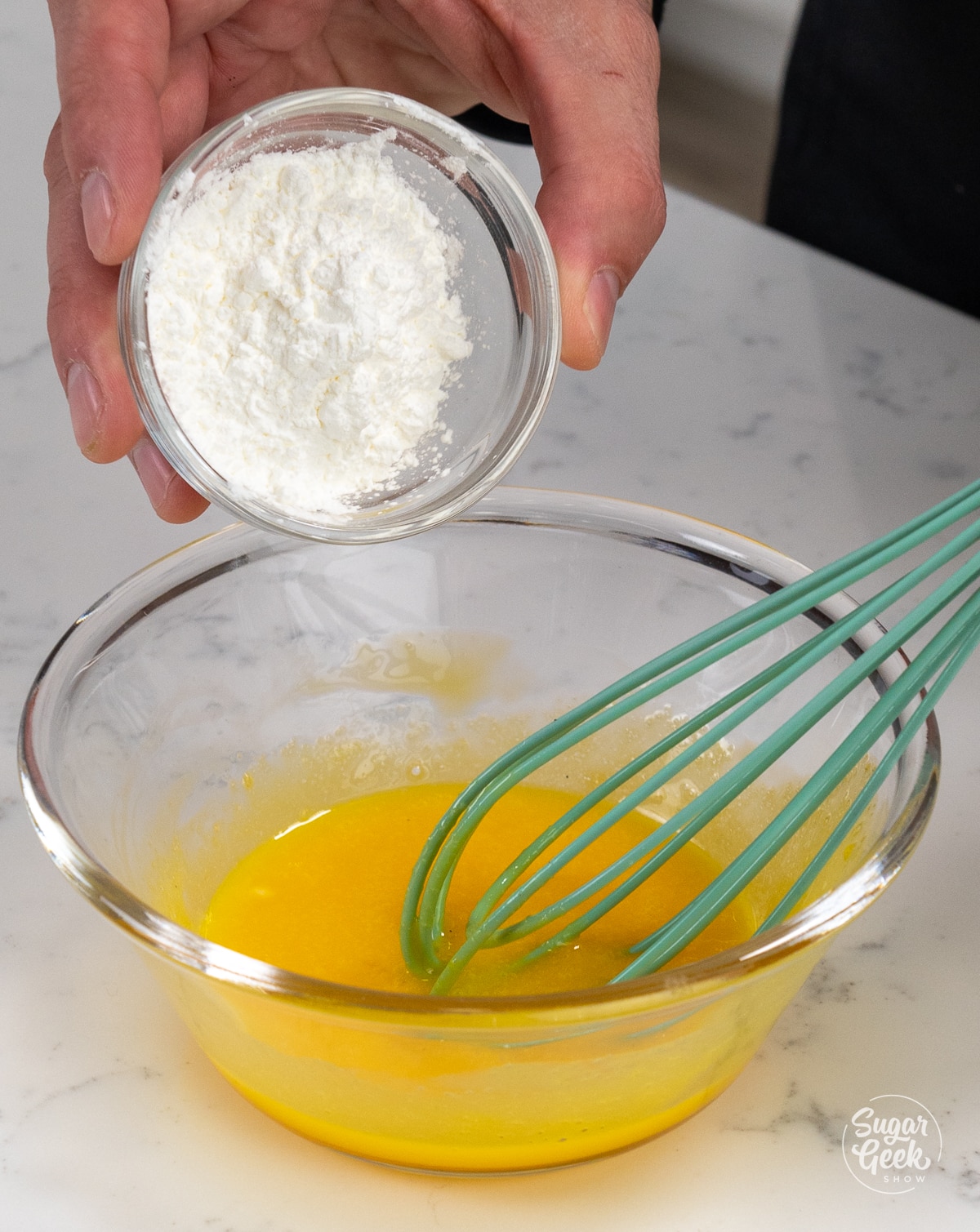 hand adding a small bowl of cornstarch to a large bowl of egg yolks