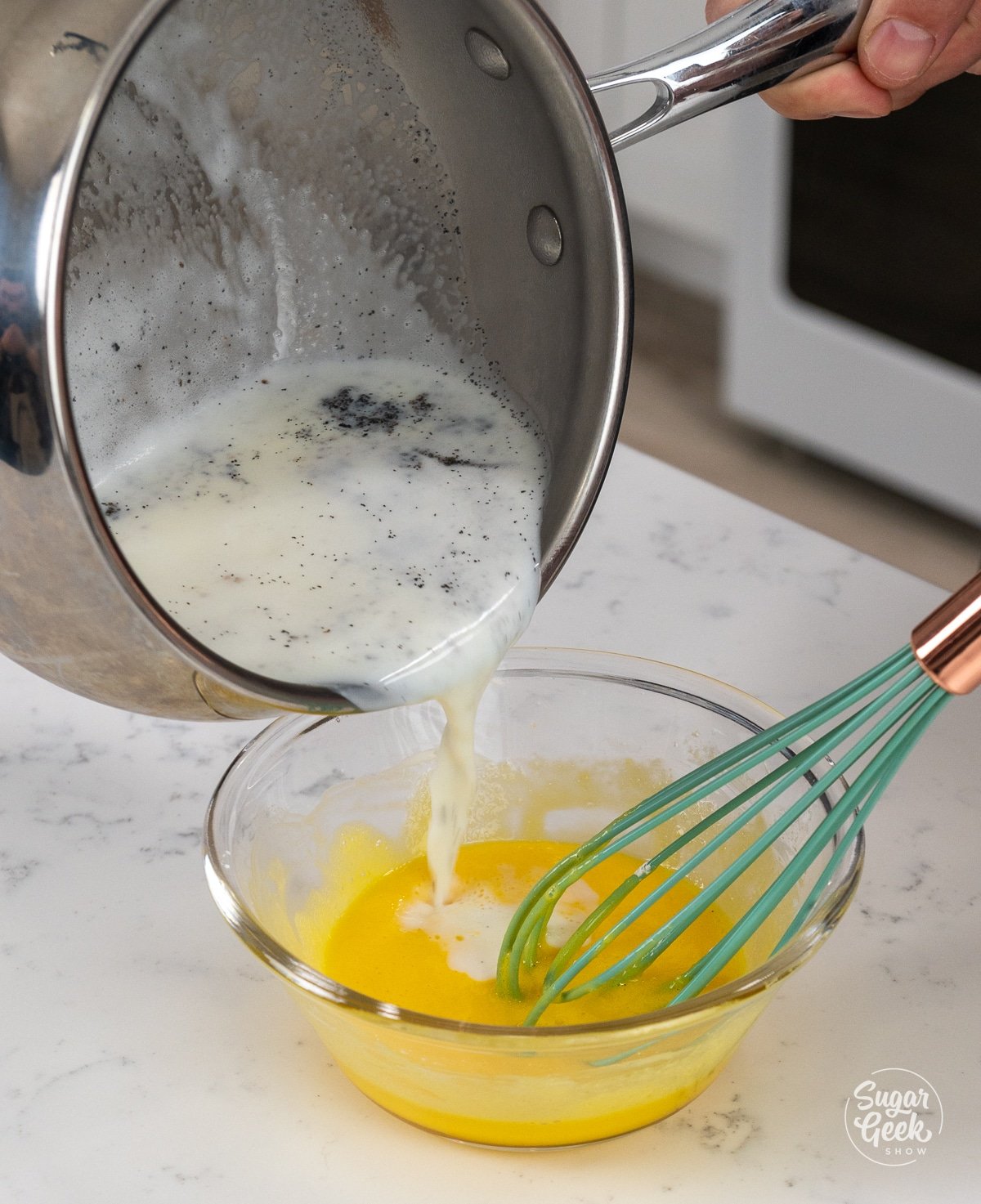 pouring a pan of hot milk into a bowl of egg yolks with a blue whisk
