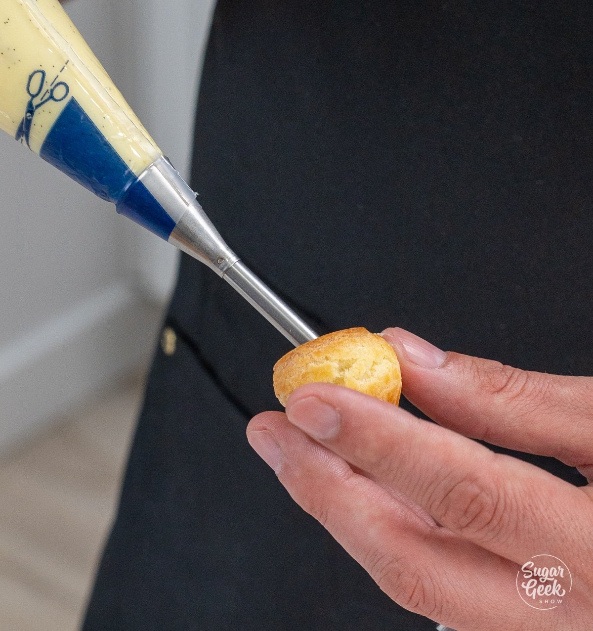Hand holding a cream puff shell while a pastry bag and long tip fills it with cream.