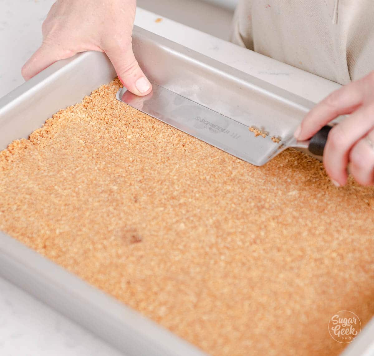 hands holding offset spatula, pressing graham cracker crust into the edges of a pan