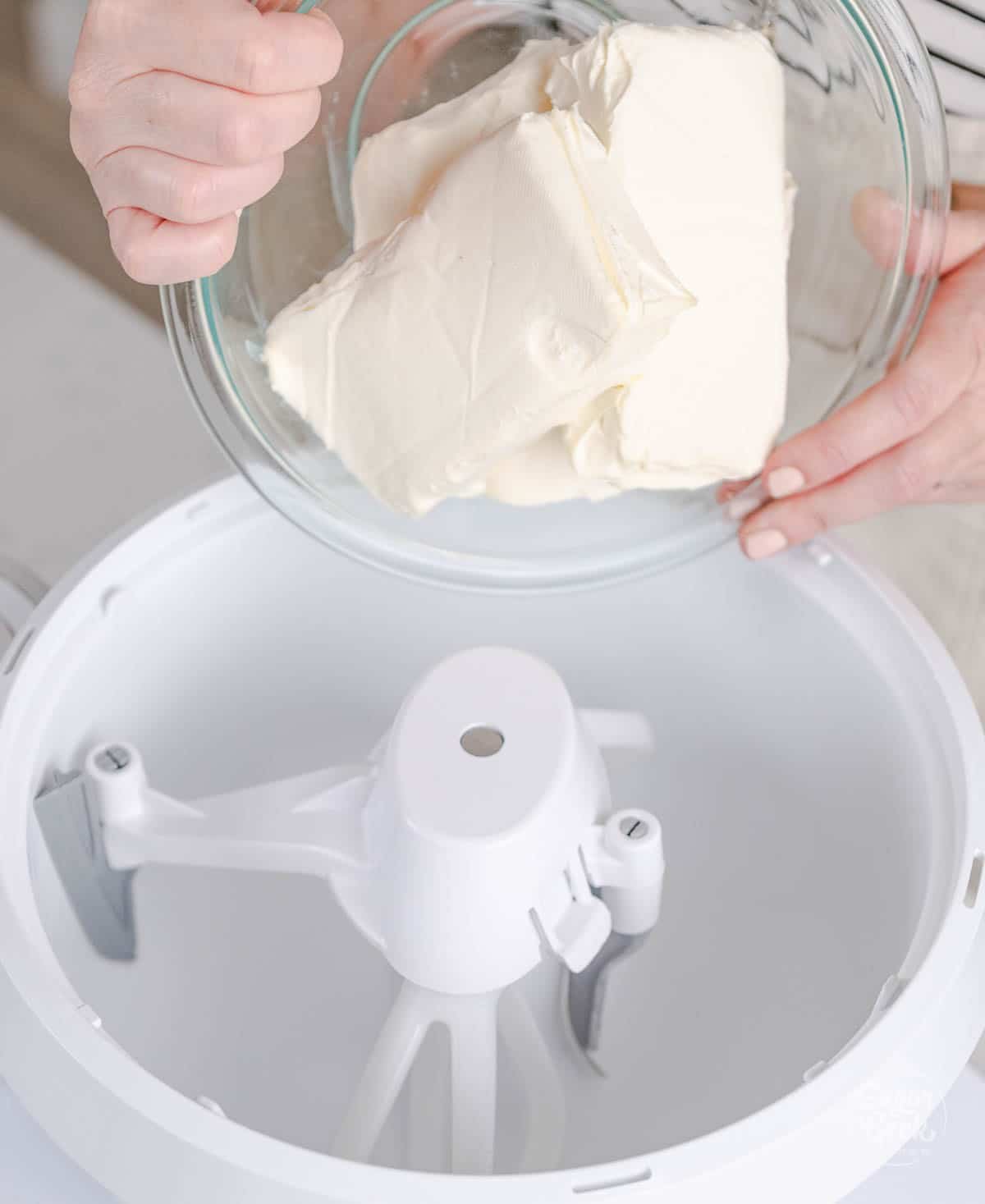 hands holding a bowl of cream cheese over a stand mixer