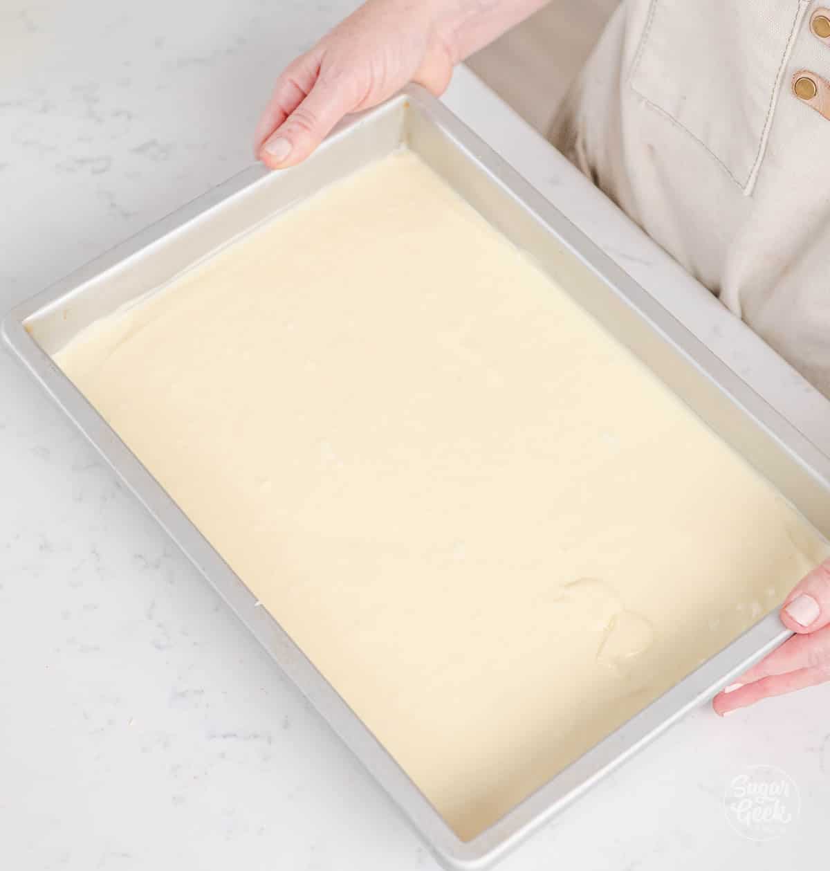 hands holding a pan full of spread out white cheesecake batter 