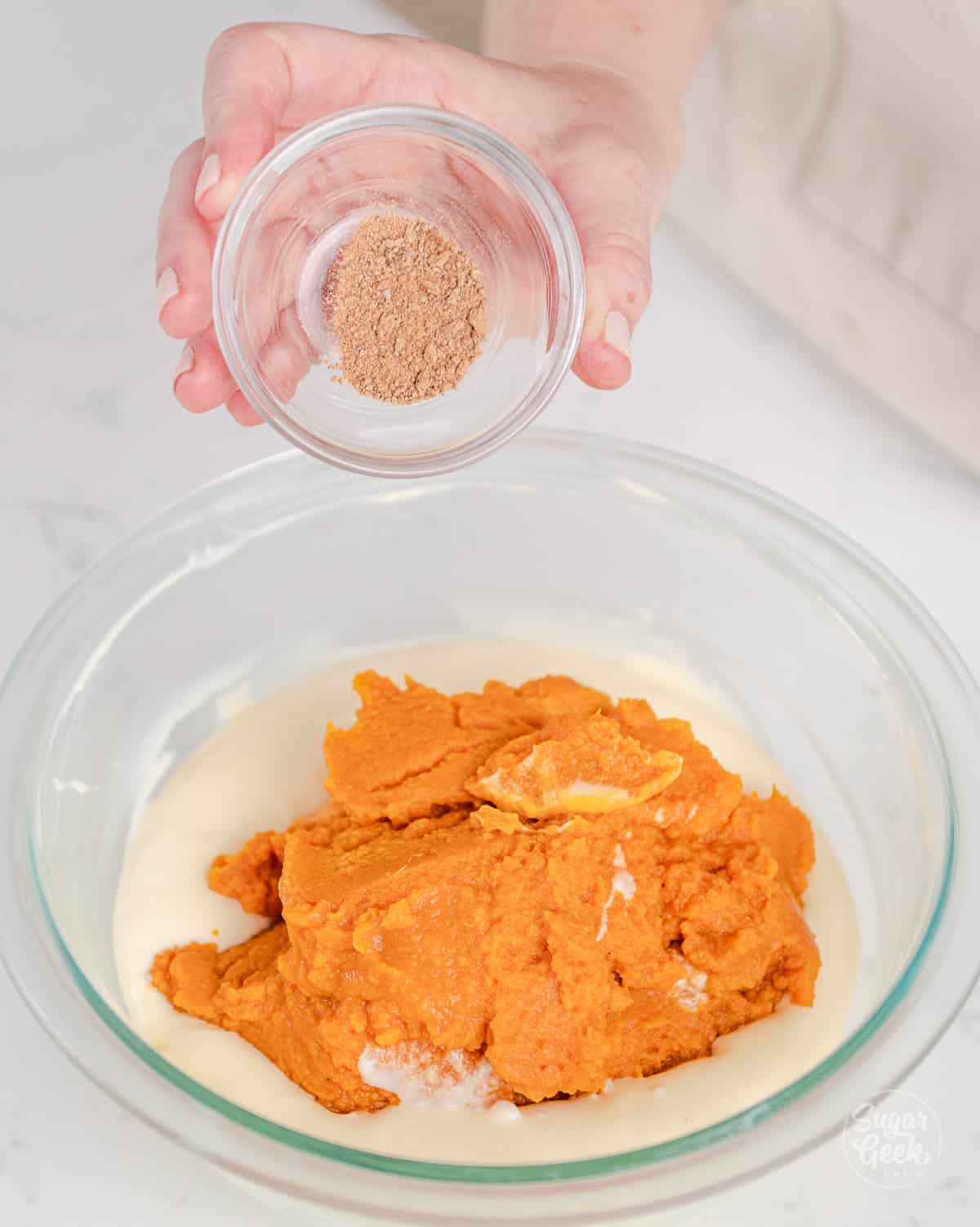 hand holding a clear bowl of nutmeg over a bowl of orange pumpkin puree