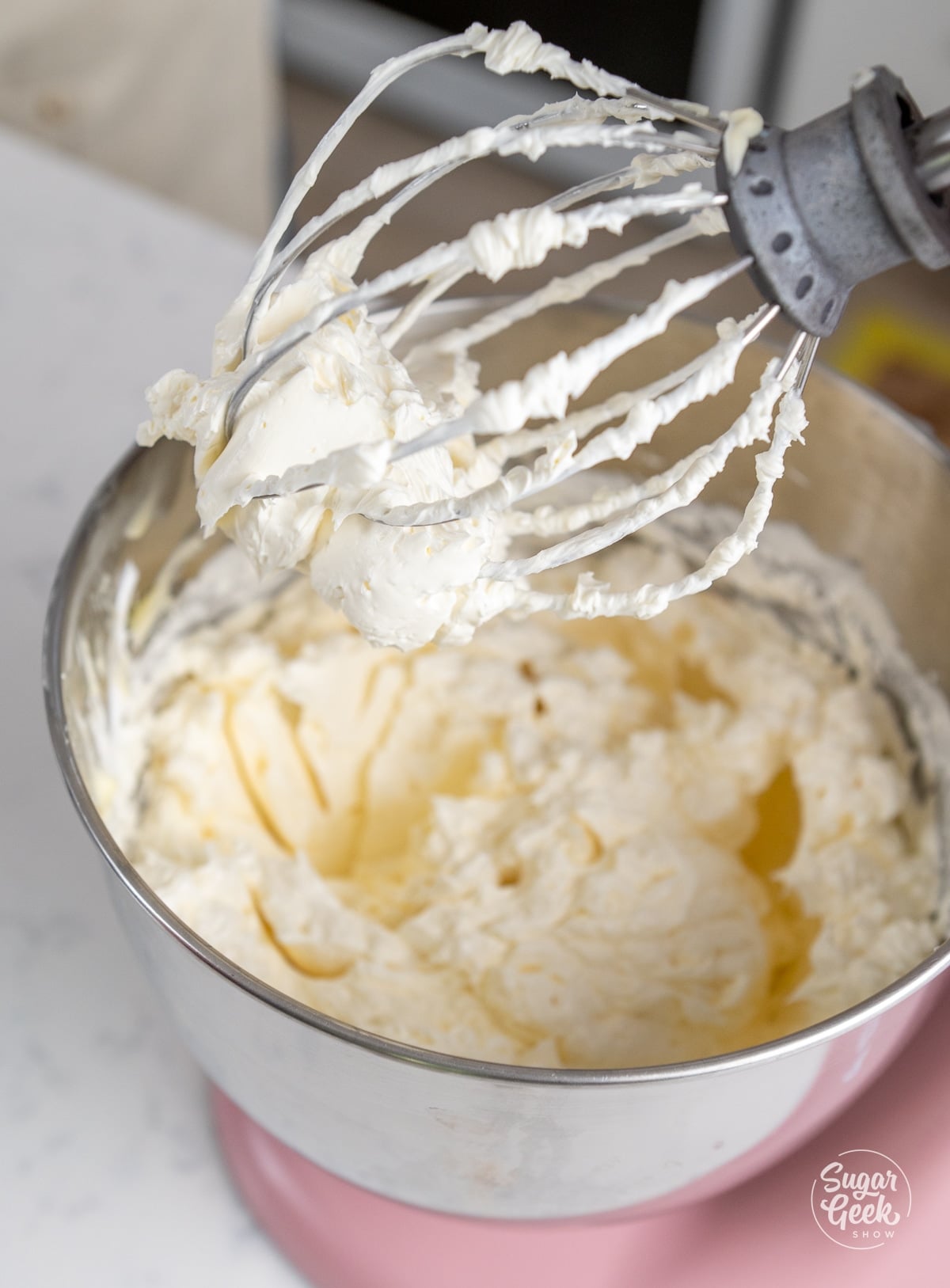 Close up of whipped swiss meringue buttercream on a whisk in the bowl of a stand mixer.