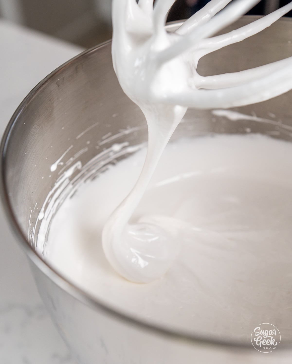 Close up of Swiss meringue on a whisk attachment above the bowl of a stand mixer.