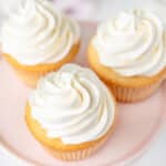 Swirls of swiss meringue buttercream on vanilla cupcakes on a pink plate.