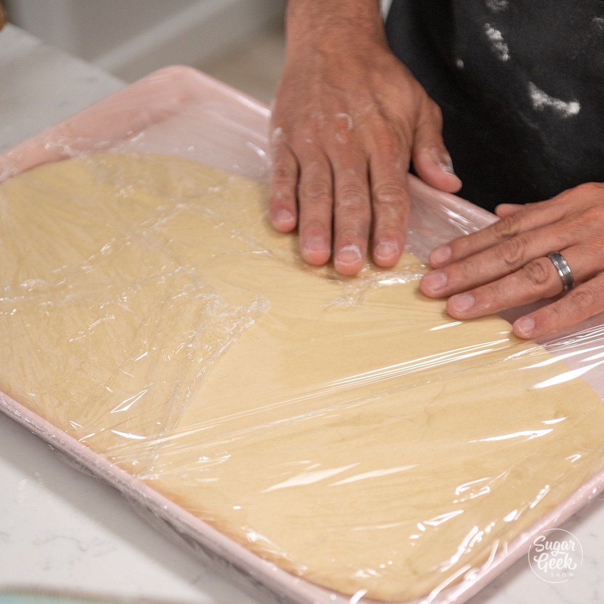 hands pressing plastic wrap over croissant dough on a sheet pan