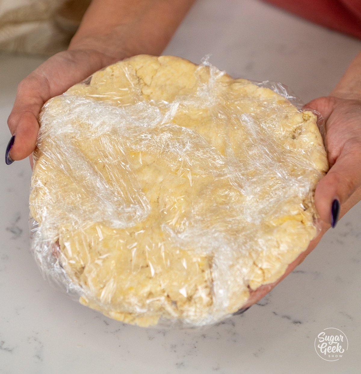 hands holding pie dough wrapped in plastic wrap. 