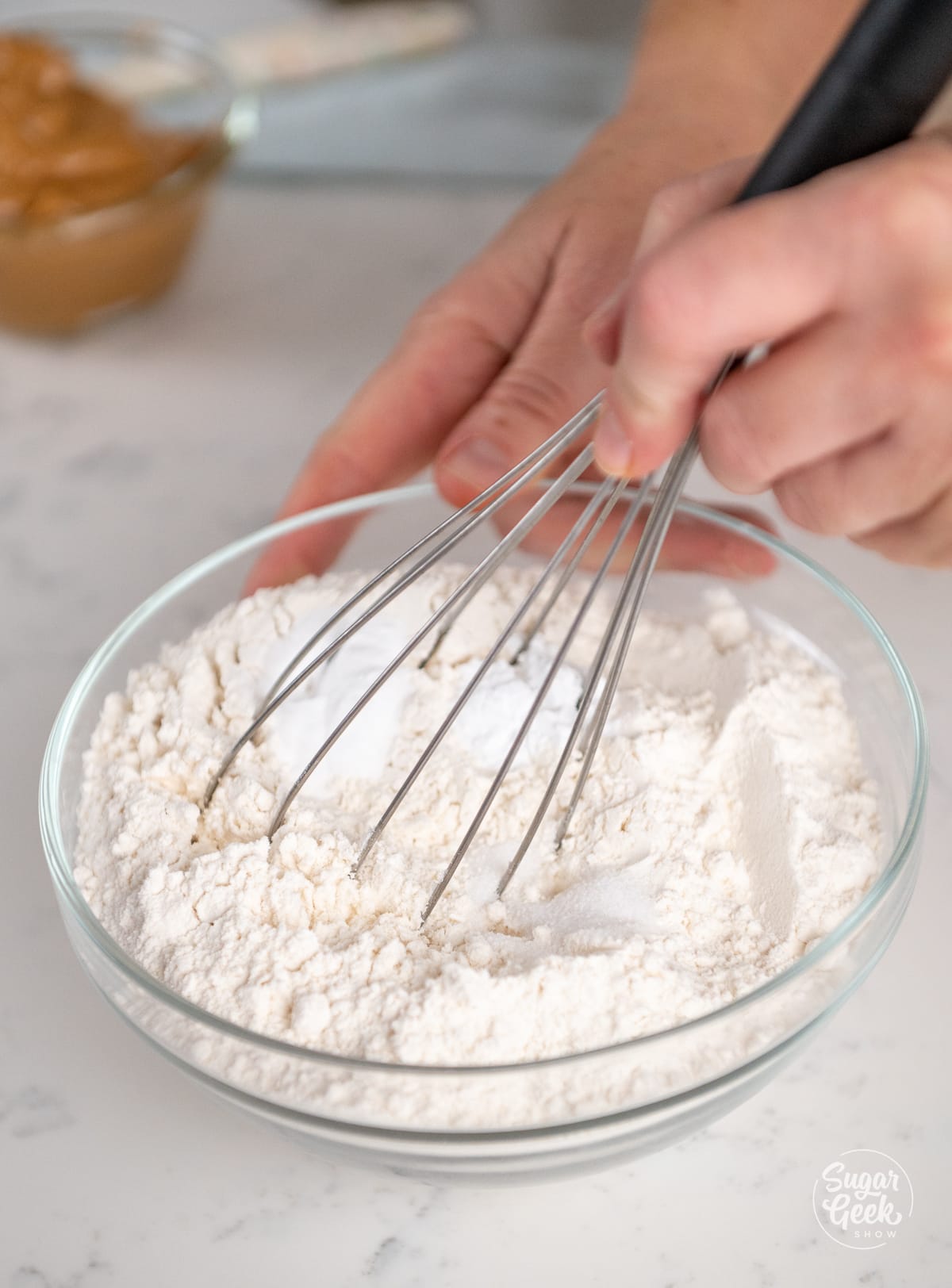 hand holding a whisk inside of a glass bowl of flour