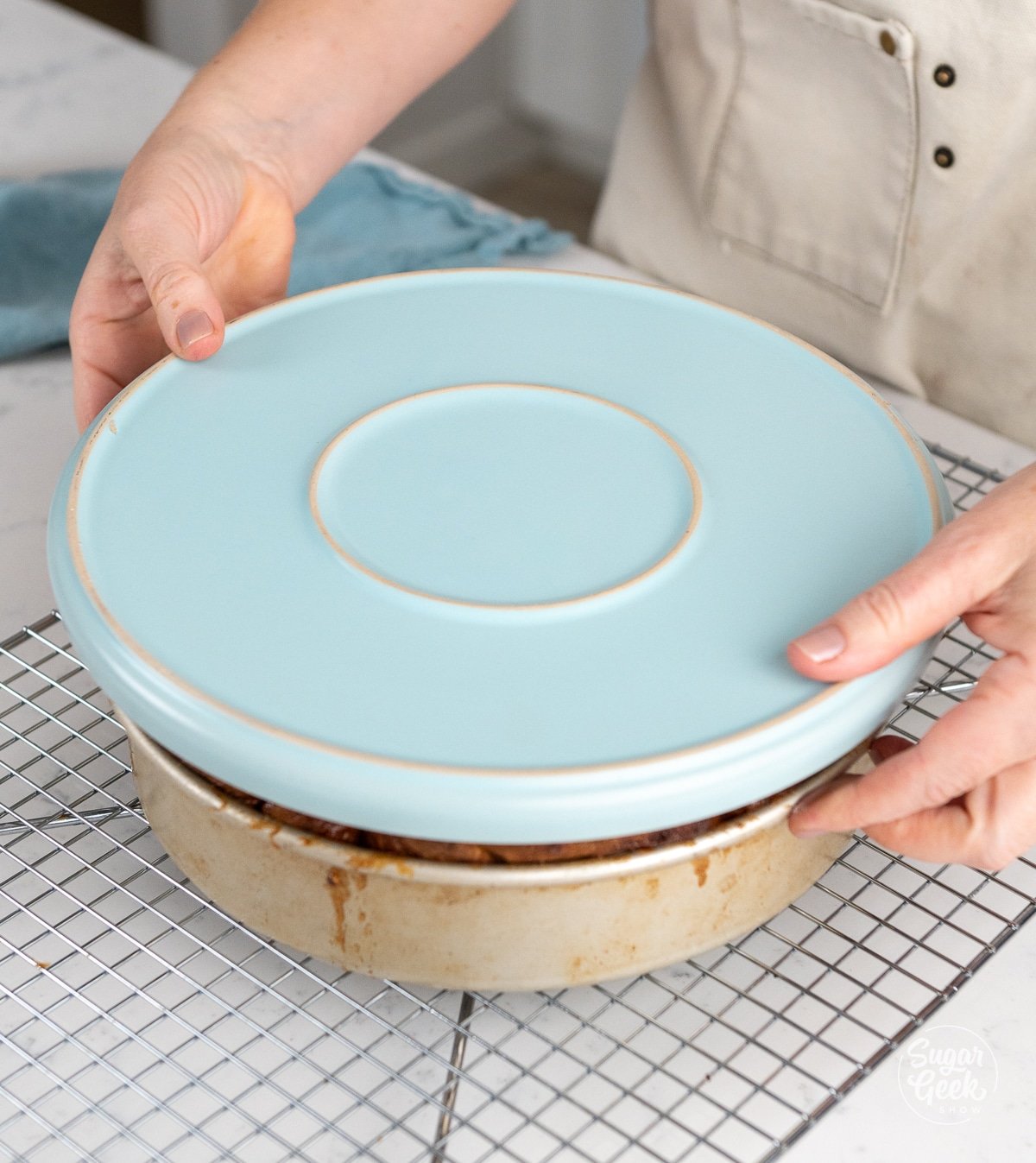 hands placing a blue plate upside down over sticky buns in a pan