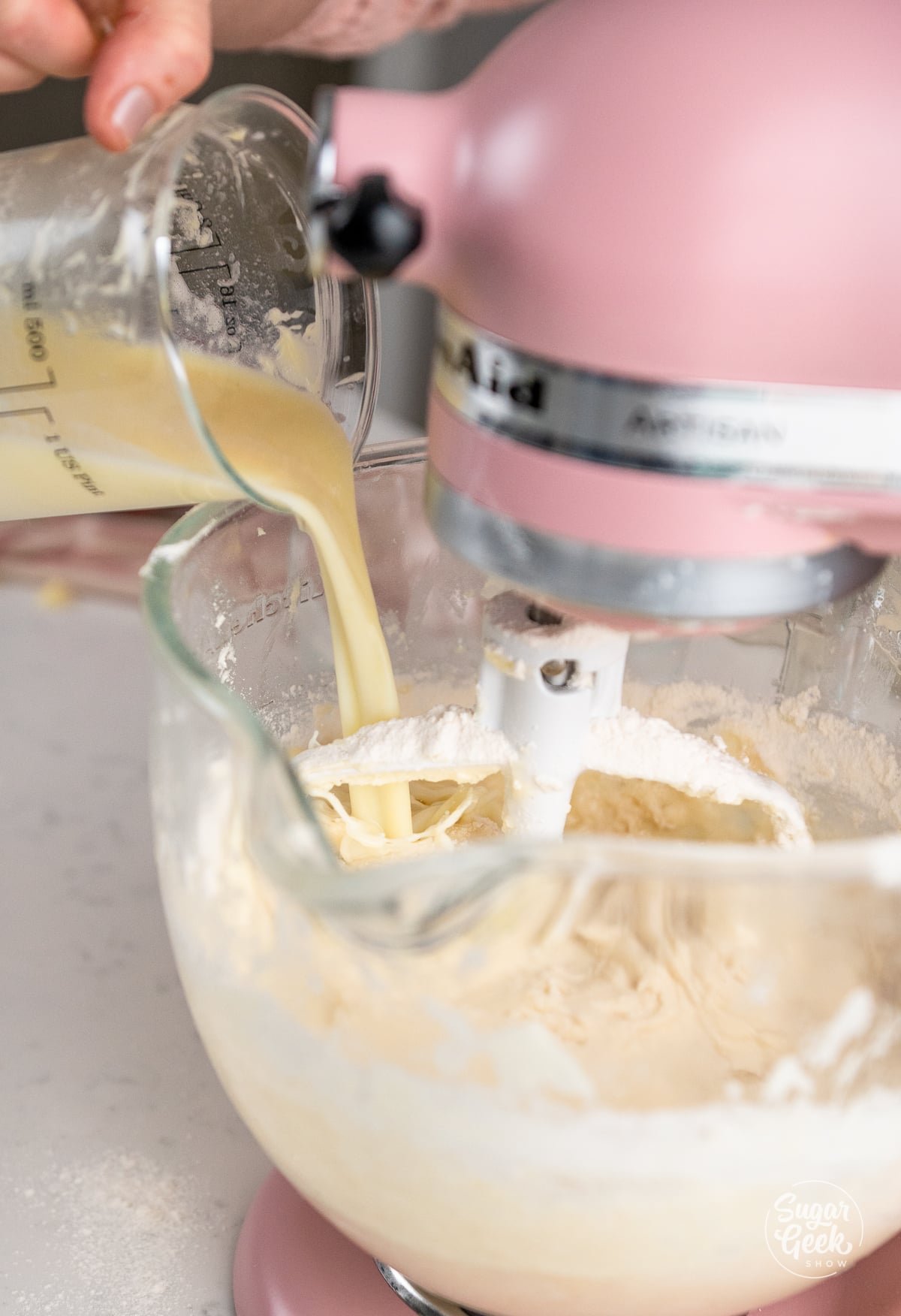 hand pouring wet ingredients into batter in glass stand mixer bowl
