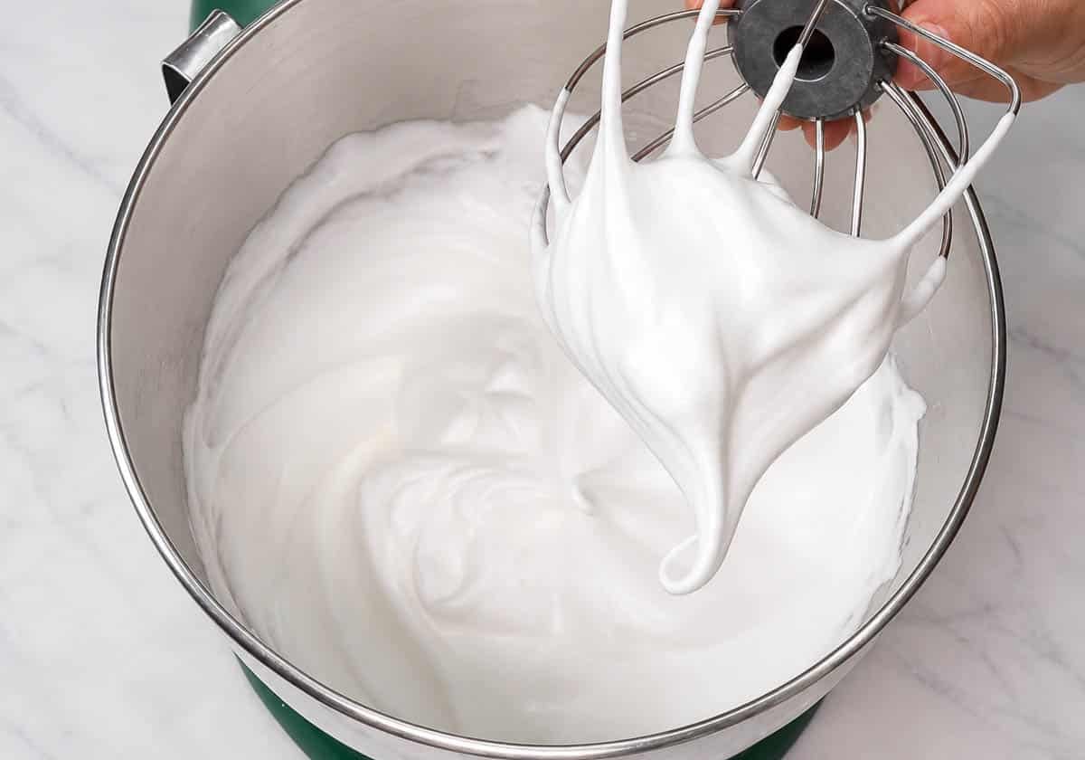 Close up of thick meringue batter on a whisk attachment over a mixing bowl.