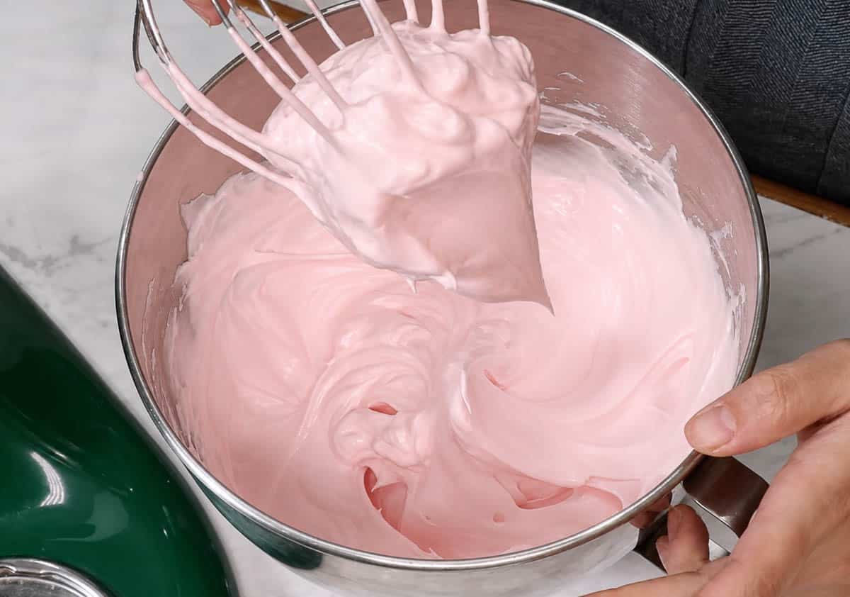 Lightly pink meringue on the whisk attachment above a mixing bowl.