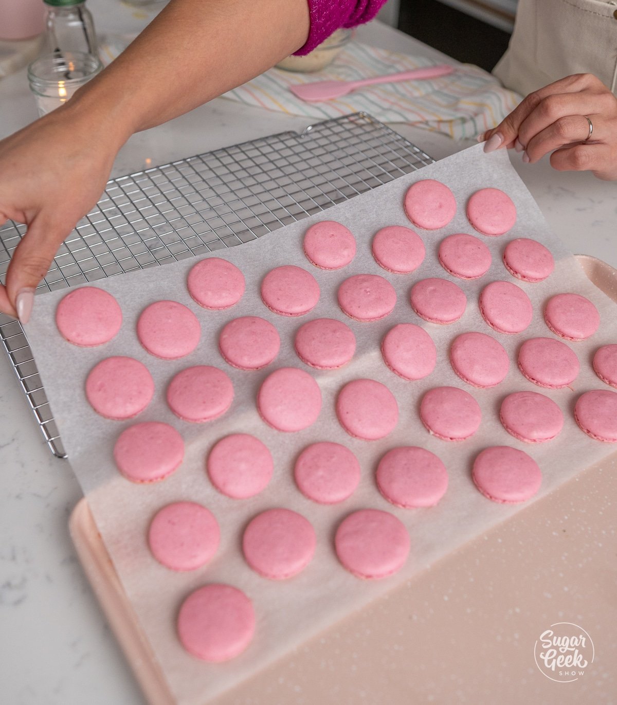 hands moving a sheet of parchment paper with baked macarons onto a wire rack
