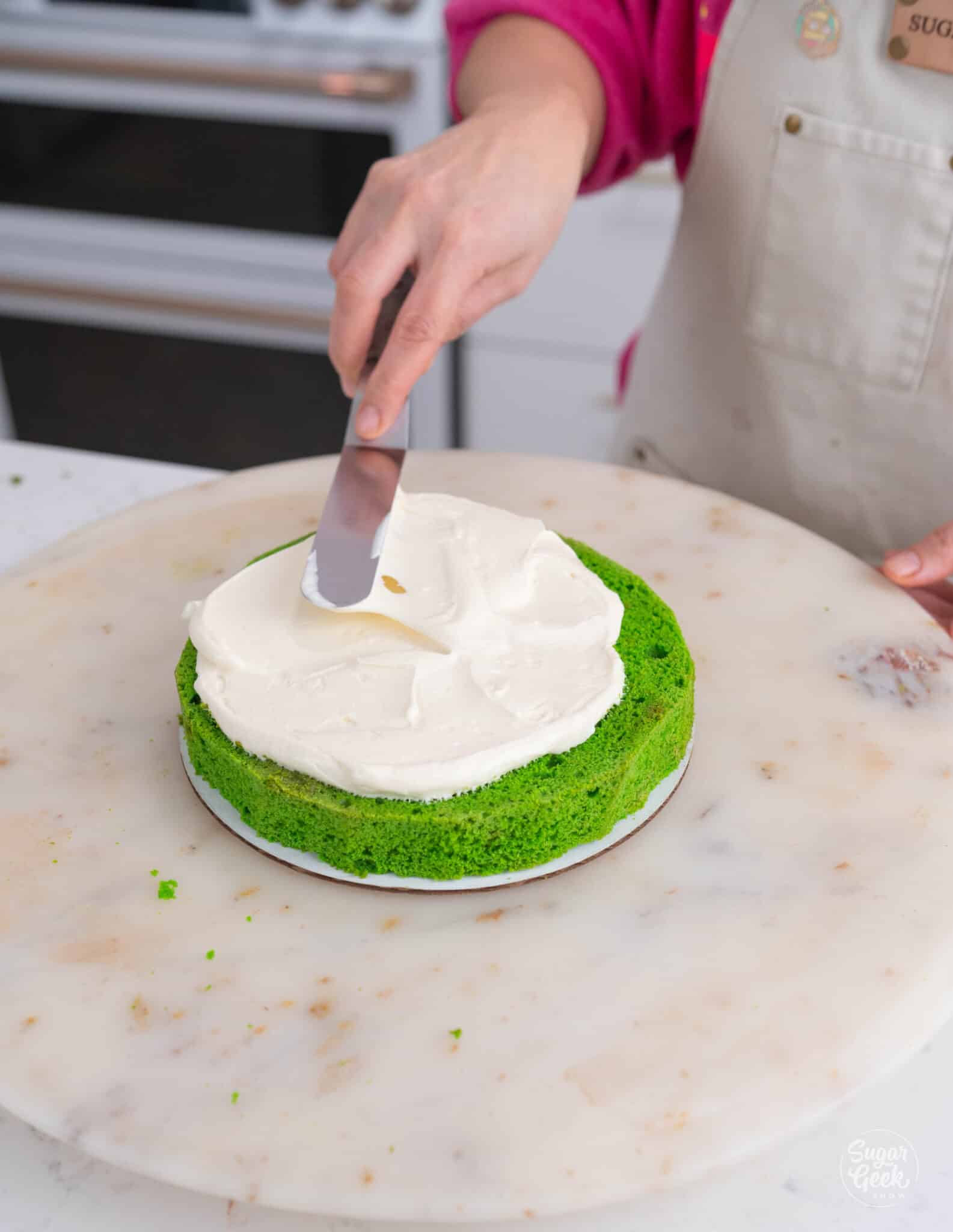 hand spreading frosting on a green cake layer with a spatula