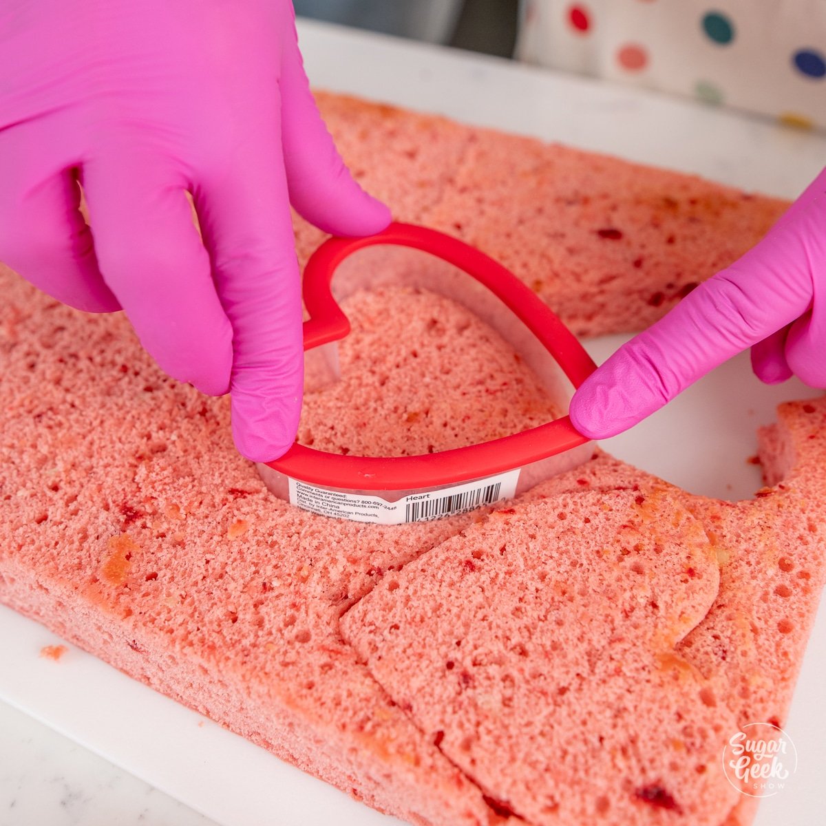 hands pushing a heart shaped cutter into a pink cake