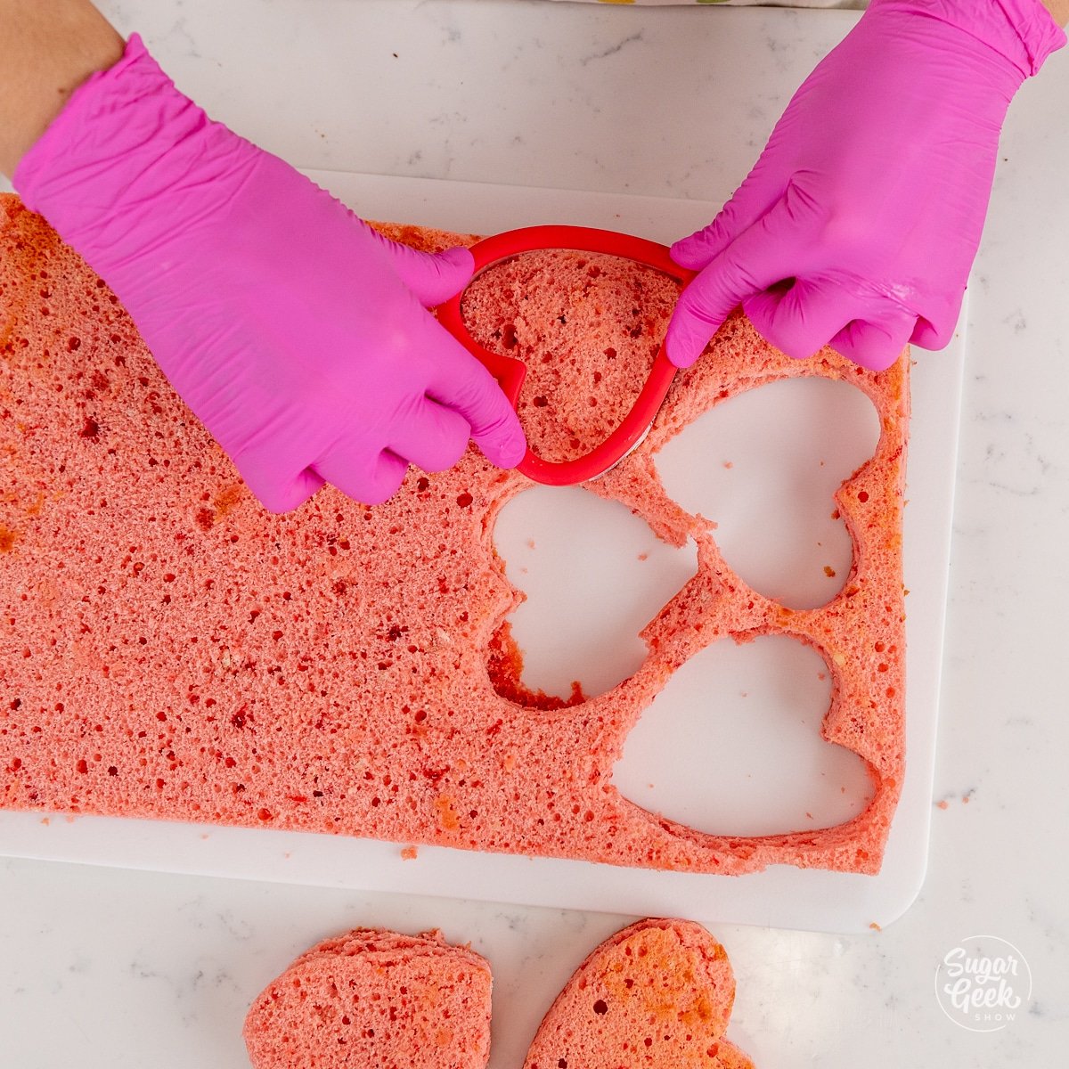 hands pushing a heart shaped cutter into a pink cake