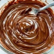 Close up of smooth and shiny chocolate ganache with a silver spoon in a glass bowl.