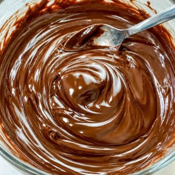 Close up of smooth and shiny chocolate ganache with a silver spoon in a glass bowl.