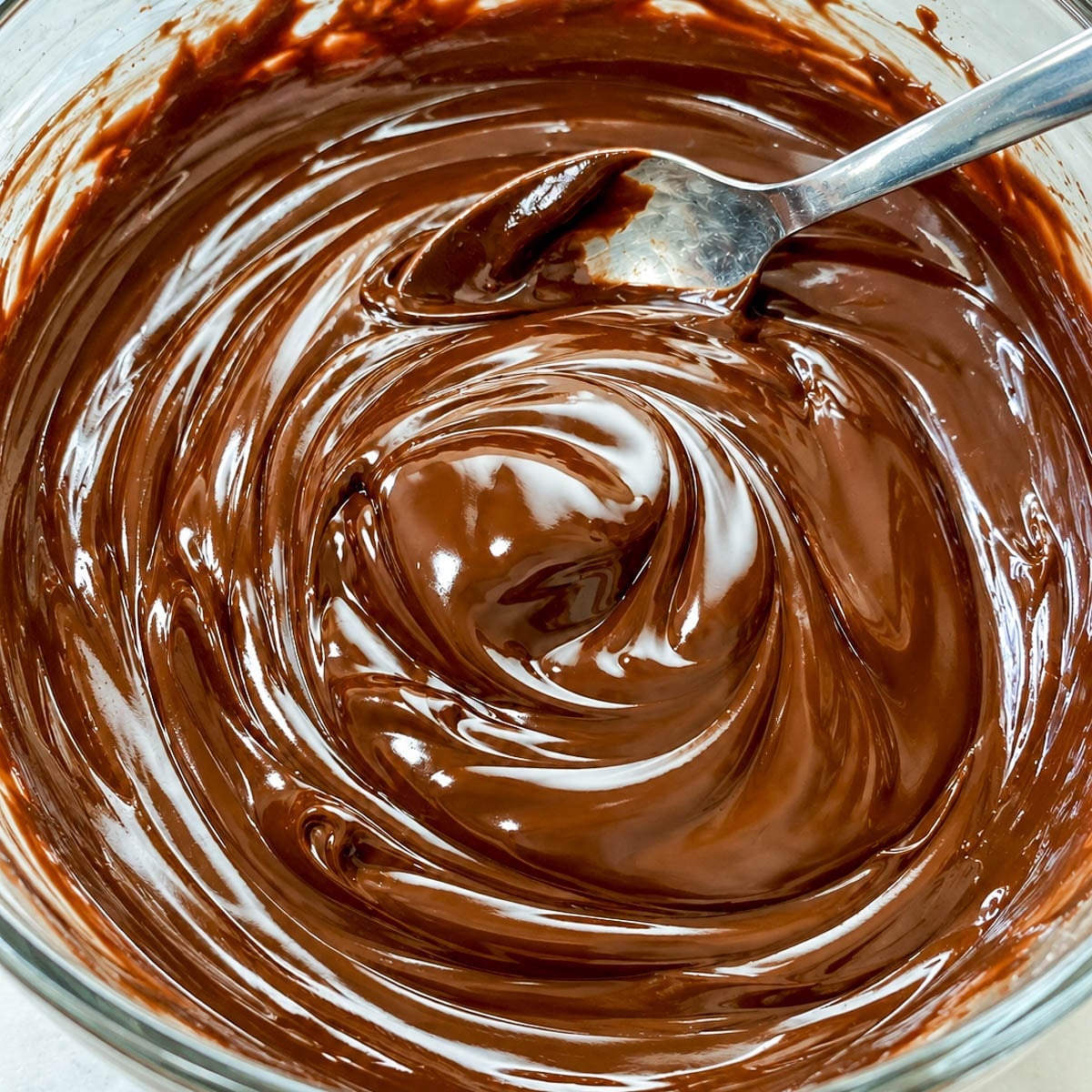 Close up of smooth and shiny chocolate ganache with a silver spoon in a glass bowl.